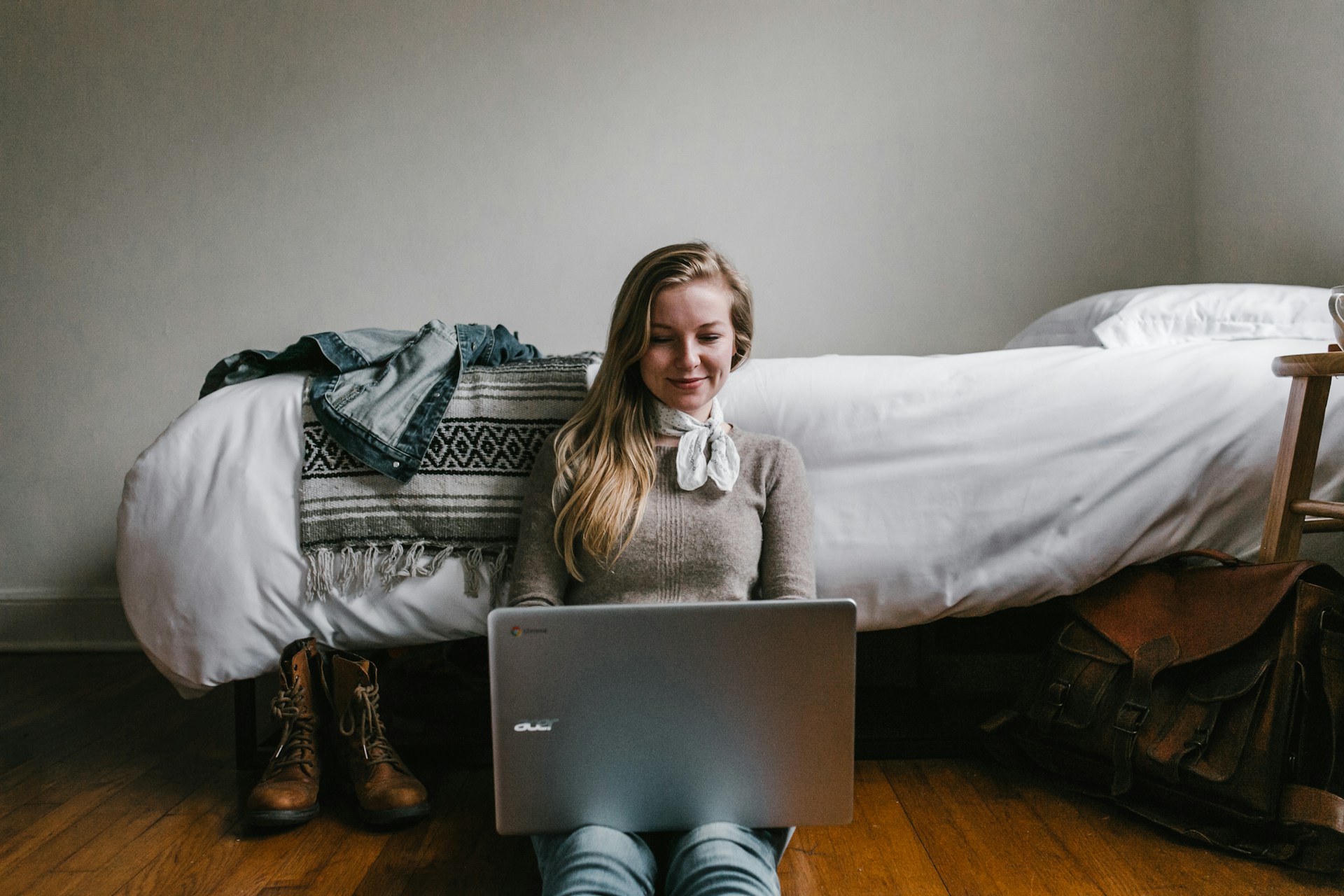 A woman is sitting on the floor near her bed and working on a laptop that is on her legs.